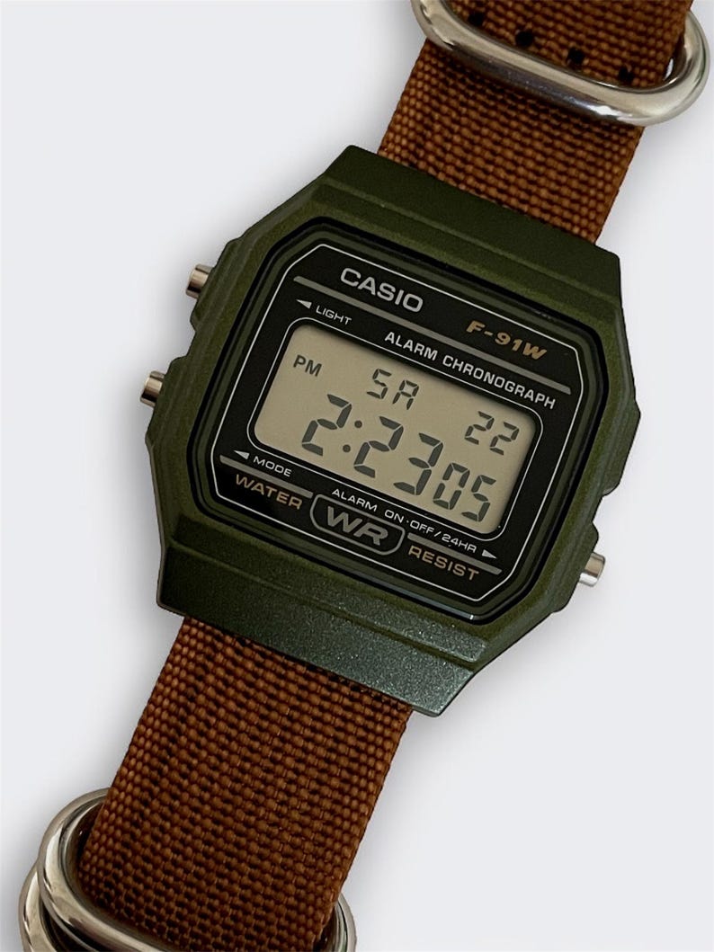 May include: A digital Casio F-91W watch with a dark green case and a brown nylon strap. The watch face displays the time as 2:23:05 PM. The watch has a light, alarm, and chronograph functions.