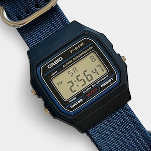 May include: A black Casio F-91W digital watch with a blue nylon strap. The watch face displays the time, day, and date. The watch has a blue and black bezel with the text "CASIO", "F-91W", and "WATER RESIST".