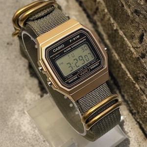 May include: A gold-toned Casio F-91W digital watch with a green fabric strap. The watch face displays the time as 3:29:07 PM. The watch has gold-colored hardware and is water-resistant.
