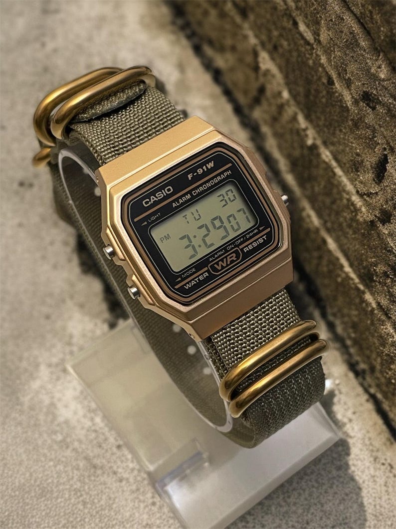 May include: A gold-toned Casio F-91W digital watch with a green fabric strap. The watch face displays the time as 3:29:07 PM. The watch has gold-colored hardware and is water-resistant.