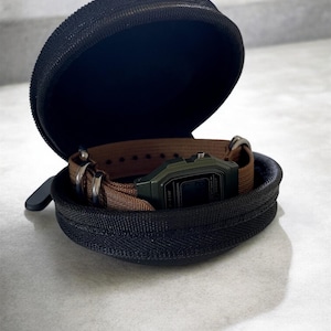 May include: A black, zipped watch case, open to reveal a digital watch with a green face and brown strap. The case is round and has a black interior. The watch is secured inside the case with the strap.