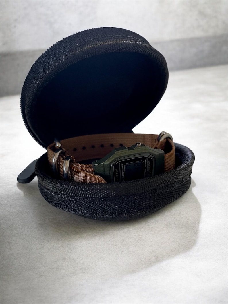 May include: A black, zipped watch case, open to reveal a digital watch with a green face and brown strap. The case is round and has a black interior. The watch is secured inside the case with the strap.