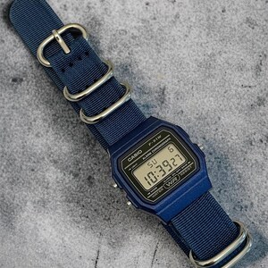 Puede incluir: Un reloj de pulsera digital azul marino con esfera rectangular y correa de nailon azul marino a juego con hebillas y anillas de metal plateado. La pantalla muestra el texto CASIO F-91W, ALARM CHRONOGRAPH y la hora 10:39:27.