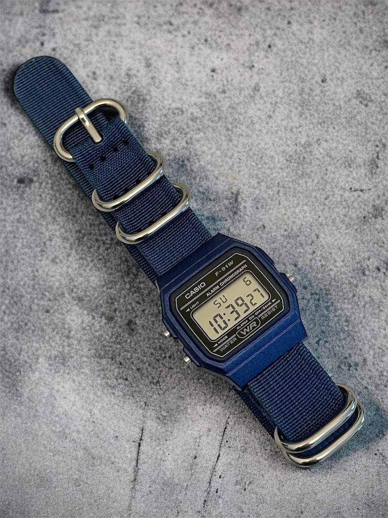 Puede incluir: Un reloj de pulsera digital azul marino con esfera rectangular y correa de nailon azul marino a juego con hebillas y anillas de metal plateado. La pantalla muestra el texto CASIO F-91W, ALARM CHRONOGRAPH y la hora 10:39:27.