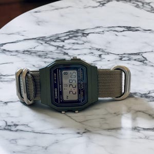 May include: An olive green Casio F-91W digital watch with a matching canvas band. The watch face displays the time, day, and date. The watch has silver-tone metal hardware and is water resistant.
