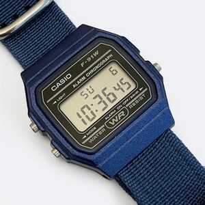 Puede incluir: Un reloj digital Casio F-91W de color azul oscuro con una correa de nailon azul a juego y herrajes de metal plateado. La esfera rectangular presenta una pantalla negra con texto blanco que dice CASIO, F-91W, ALARM CHRONOGRAPH y WATER RESIST.