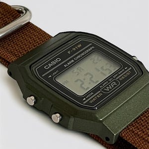 May include: A close-up of a Casio F-91W digital watch with a green case and a brown nylon strap. The watch face displays the time and has the text "CASIO" and "F-91W". The watch is water resistant.