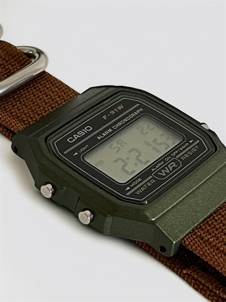 May include: A close-up of a Casio F-91W digital watch with a green case and a brown nylon strap. The watch face displays the time and has the text "CASIO" and "F-91W". The watch is water resistant.