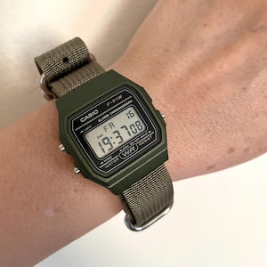 May include: A digital Casio F-91W watch with a green case and a khaki fabric strap. The watch face displays the time, day, and date. The watch is on a wrist.