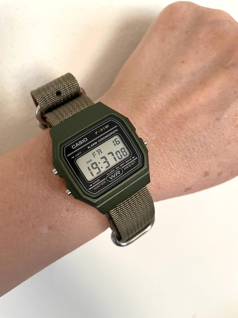May include: A digital Casio F-91W watch with a green case and a khaki fabric strap. The watch face displays the time, day, and date. The watch is on a wrist.