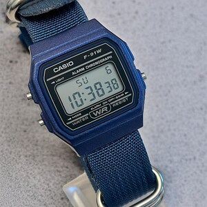 Puede incluir: Un reloj de pulsera digital azul con caja rectangular y correa de nailon tejido azul a juego con herrajes de metal plateado. La esfera muestra el texto CASIO F-91W, ALARM CHRONOGRAPH y WATER WR RESIST, junto con una lectura de hora digital.