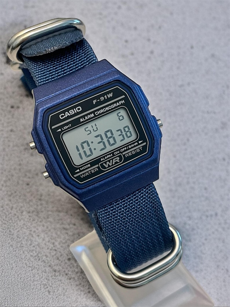 Puede incluir: Un reloj de pulsera digital azul con caja rectangular y correa de nailon tejido azul a juego con herrajes de metal plateado. La esfera muestra el texto CASIO F-91W, ALARM CHRONOGRAPH y WATER WR RESIST, junto con una lectura de hora digital.