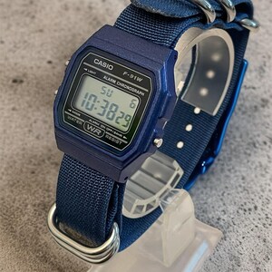 Puede incluir: Un reloj de pulsera digital azul con caja rectangular y el texto CASIO F-91W ALARM CHRONOGRAPH WATER RESIST impreso en la esfera. El reloj tiene una correa de nailon azul con hebillas de metal plateado y una pantalla digital que muestra 10:3829.