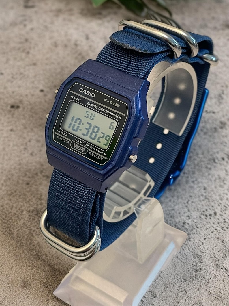 Puede incluir: Un reloj de pulsera digital azul con caja rectangular y el texto CASIO F-91W ALARM CHRONOGRAPH WATER RESIST impreso en la esfera. El reloj tiene una correa de nailon azul con hebillas de metal plateado y una pantalla digital que muestra 10:3829.