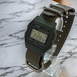 May include: A digital watch with a dark green case and a matching green nylon strap. The watch face displays the time, day, and date. The watch is labeled "CASIO F-91W" and has silver-tone metal hardware.