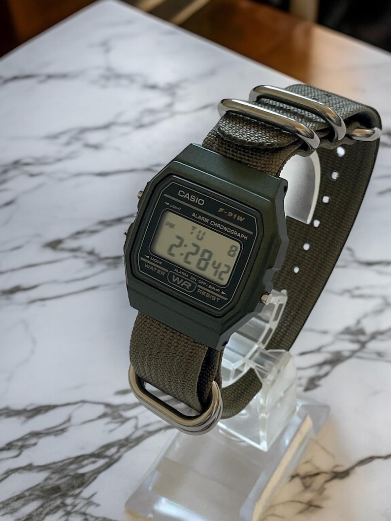 May include: A digital watch with a dark green case and a matching green nylon strap. The watch face displays the time, day, and date. The watch is labeled "CASIO F-91W" and has silver-tone metal hardware.