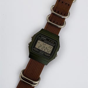 May include: A digital Casio F-91W watch with a dark green case and a brown nylon strap. The watch face displays the time as 2:22:52. The strap has silver-coloured metal loops and a buckle.