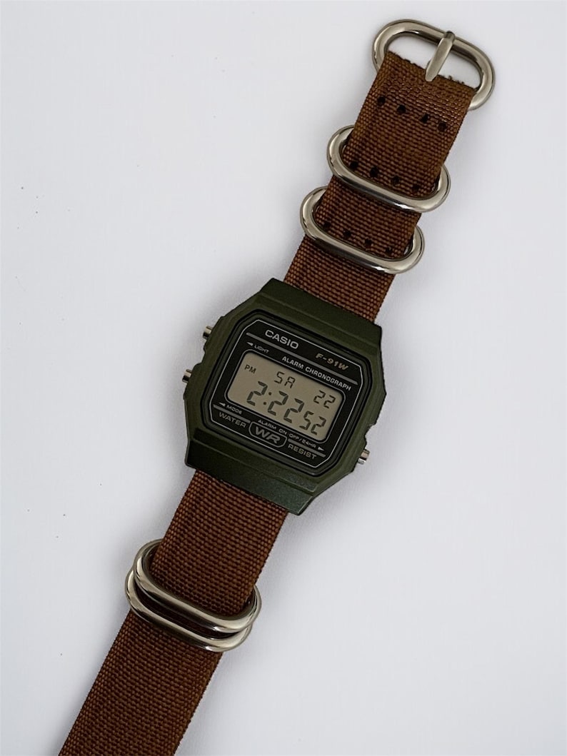 May include: A digital Casio F-91W watch with a dark green case and a brown nylon strap. The watch face displays the time as 2:22:52. The strap has silver-coloured metal loops and a buckle.