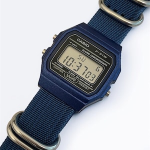 Puede incluir: Un reloj de pulsera digital azul oscuro con caja rectangular y correa de nailon tejido a juego con anillas met&aacute;licas plateadas. La esfera muestra el texto CASIO, F-91W, ALARM CHRONOGRAPH y WATER RESIST, junto a una lectura digital.