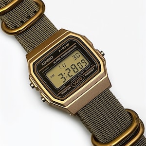 May include: A gold-toned Casio F-91W digital watch with a green fabric band. The watch face displays the time and date. The band features gold-colored metal loops. The watch is water-resistant.