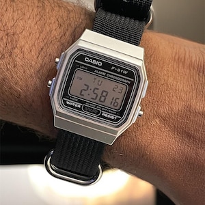 May include: A silver Casio F-91W digital watch with a black nylon strap. The watch face displays the time, date, and day of the week. The watch is water resistant and has an alarm and chronograph functions.