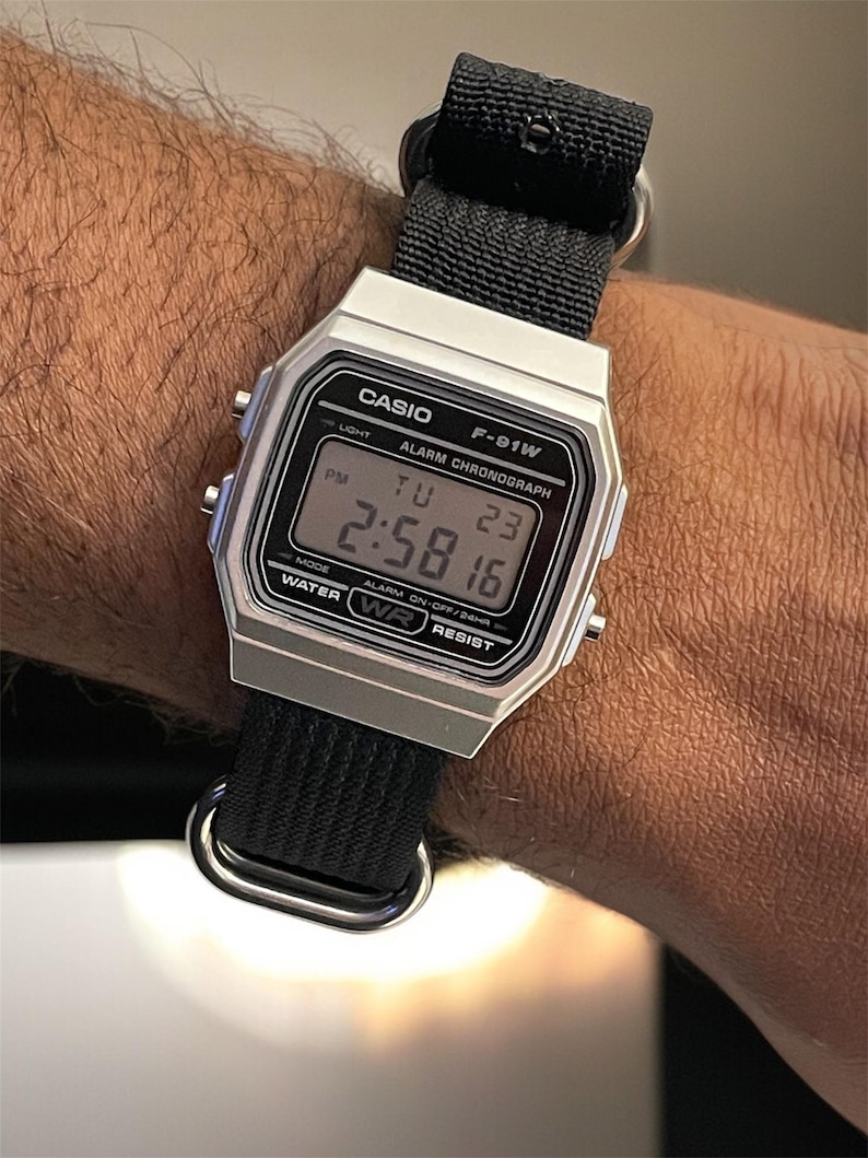 May include: A silver Casio F-91W digital watch with a black nylon strap. The watch face displays the time, date, and day of the week. The watch is water resistant and has an alarm and chronograph functions.