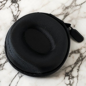 May include: A black, circular watch case with a textured surface. It features a zip closure and an internal, oval-shaped cushion for securing a watch. The background displays a white and grey marble pattern.