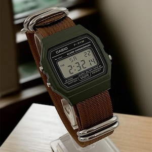 May include: A Casio F-91W digital watch with a green case and brown fabric strap. The watch displays the time as 2:32 PM. The watch is on a clear display stand.