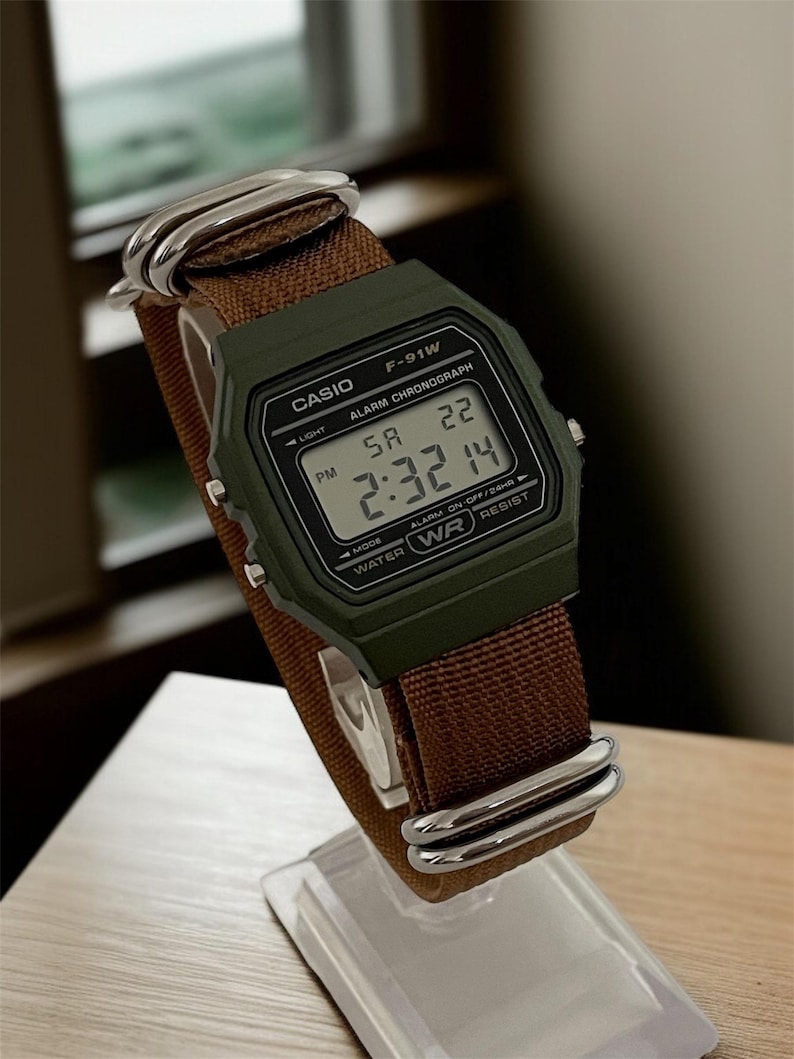 May include: A Casio F-91W digital watch with a green case and brown fabric strap. The watch displays the time as 2:32 PM. The watch is on a clear display stand.