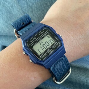 Puede incluir: Un reloj digital Casio F-91W azul con una correa de tela azul a juego. La esfera del reloj muestra el texto CASIO, F-91W, ALARM CHRONOGRAPH y WATER WR RESIST. La pantalla digital muestra la hora 11:00:39, el d&iacute;a SU y la fecha 6.