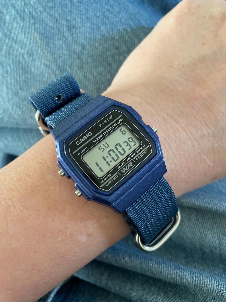 Puede incluir: Un reloj digital Casio F-91W azul con una correa de tela azul a juego. La esfera del reloj muestra el texto CASIO, F-91W, ALARM CHRONOGRAPH y WATER WR RESIST. La pantalla digital muestra la hora 11:00:39, el d&iacute;a SU y la fecha 6.