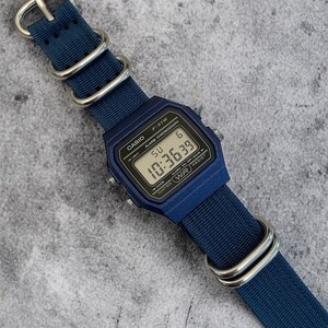 Puede incluir: Un reloj digital azul marino con caja rectangular y correa de nailon azul marino a juego con hebillas de metal plateado. La esfera muestra el texto CASIO F-91W, ALARM CHRONOGRAPH y WATER WR RESIST, con la hora digital de 10:36:39.
