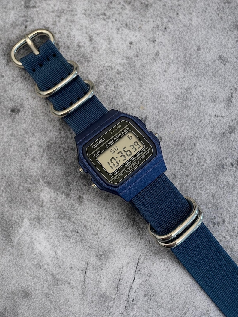 Puede incluir: Un reloj digital azul marino con caja rectangular y correa de nailon azul marino a juego con hebillas de metal plateado. La esfera muestra el texto CASIO F-91W, ALARM CHRONOGRAPH y WATER WR RESIST, con la hora digital de 10:36:39.