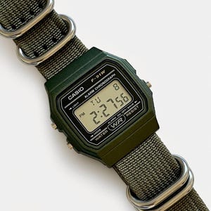 May include: A dark green Casio F-91W digital watch with a matching green nylon strap. The watch face displays the time, day, and date. The strap is secured with silver-colored metal loops.