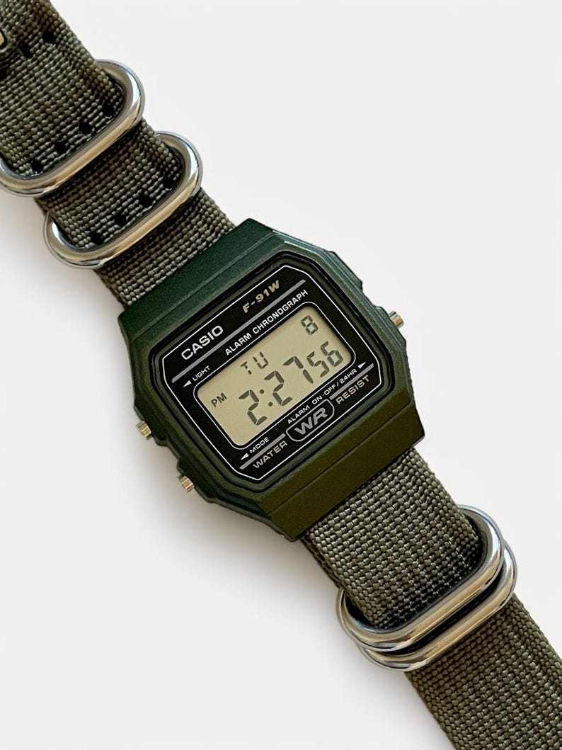 May include: A dark green Casio F-91W digital watch with a matching green nylon strap. The watch face displays the time, day, and date. The strap is secured with silver-colored metal loops.