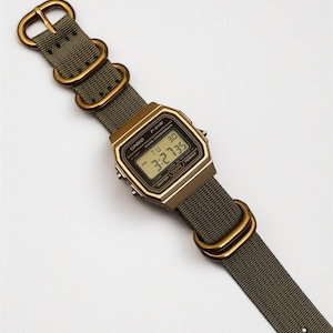 May include: A digital Casio F-91W watch with a gold-tone case and olive green nylon strap. The watch face displays the time, day, and date. The strap features gold-colored metal hardware.