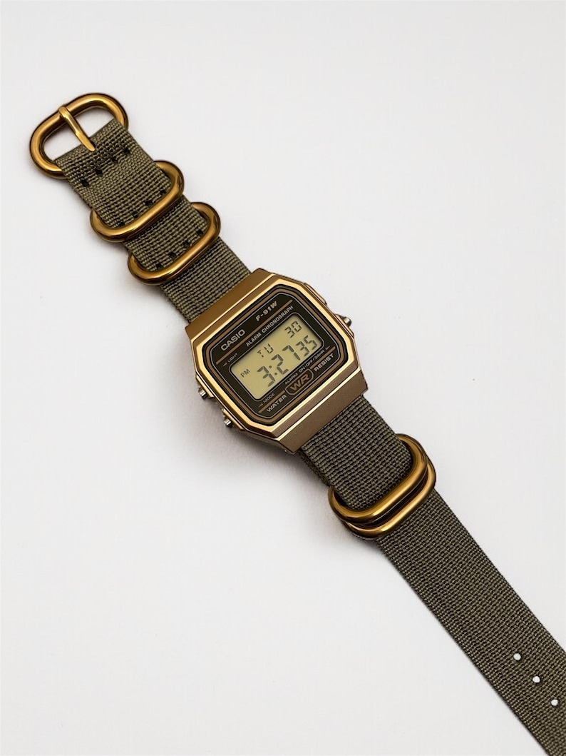May include: A digital Casio F-91W watch with a gold-tone case and olive green nylon strap. The watch face displays the time, day, and date. The strap features gold-colored metal hardware.