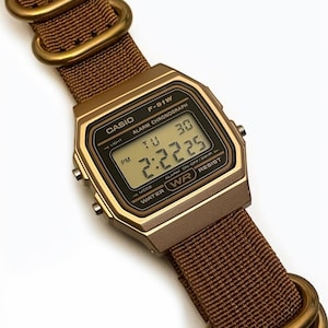 Może przedstawiać: Cyfrowy zegarek Casio F-91W z brązowym nylonowym paskiem i złotymi metalowymi elementami. Tarcza zegarka wyświetla godzinę i dzień. Zegarek ma złotą obudowę i jest wodoodporny.