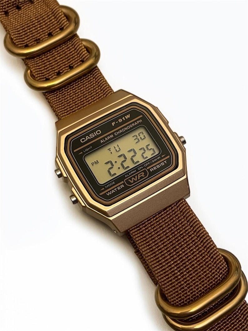 Może przedstawiać: Cyfrowy zegarek Casio F-91W z brązowym nylonowym paskiem i złotymi metalowymi elementami. Tarcza zegarka wyświetla godzinę i dzień. Zegarek ma złotą obudowę i jest wodoodporny.