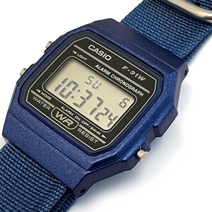 Puede incluir: Un reloj digital azul con una correa de nailon azul a juego y una hebilla de metal plateado. La esfera rectangular tiene una pantalla negra con texto blanco que dice CASIO, F-91W, ALARM CHRONOGRAPH y WATER RESIST, mostrando la hora 10:37 y el d&iacute;a SU.