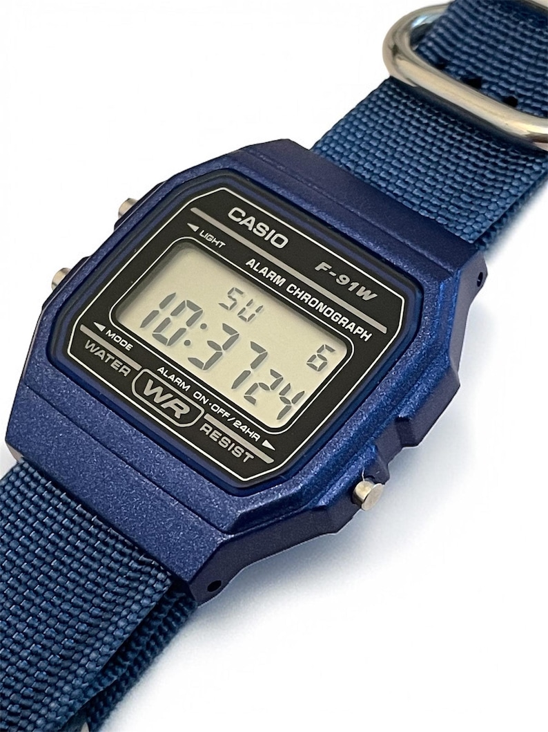 Puede incluir: Un reloj digital azul con una correa de nailon azul a juego y una hebilla de metal plateado. La esfera rectangular tiene una pantalla negra con texto blanco que dice CASIO, F-91W, ALARM CHRONOGRAPH y WATER RESIST, mostrando la hora 10:37 y el d&iacute;a SU.