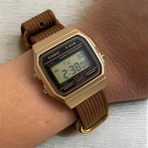Może przedstawiać: Złoty zegarek cyfrowy Casio F-91W z brązowym nylonowym paskiem. Tarcza zegarka wyświetla godzinę, dzień i datę. Zegarek jest na nadgarstku, prezentując jego design i szczeg&oacute;ły.