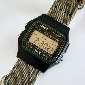 May include: A black Casio F-91W digital watch with a rectangular face, green border, and a textured olive green nylon strap featuring silver-tone metal rings. The digital display shows the time 2:30:22, the day TU, and the text ALARM CHRONOGRAPH.