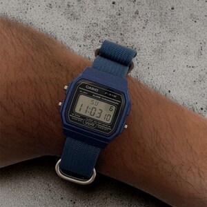 Puede incluir: Un reloj de pulsera digital azul con esfera rectangular y correa de tela azul a juego. La pantalla muestra el texto CASIO F-91W, ALARM CHRONOGRAPH, WATER RESIST, la hora 11:03 y el d&iacute;a SU 6. Cuenta con herrajes de metal de color plateado.
