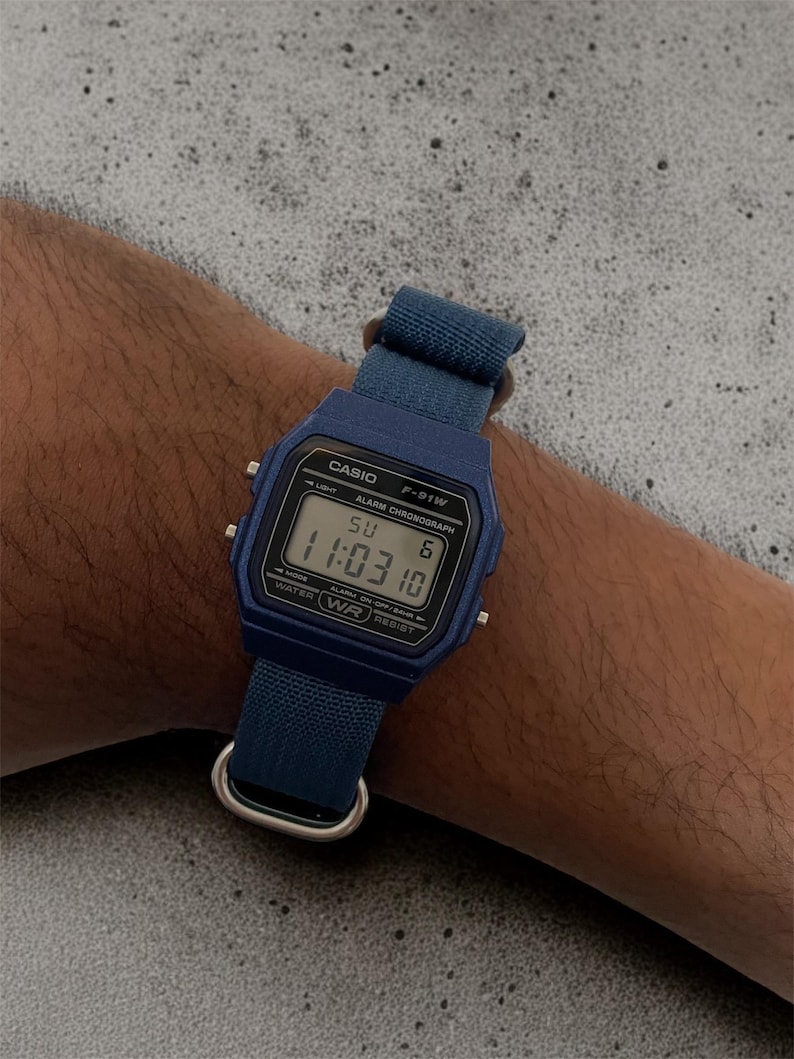 Puede incluir: Un reloj de pulsera digital azul con esfera rectangular y correa de tela azul a juego. La pantalla muestra el texto CASIO F-91W, ALARM CHRONOGRAPH, WATER RESIST, la hora 11:03 y el d&iacute;a SU 6. Cuenta con herrajes de metal de color plateado.