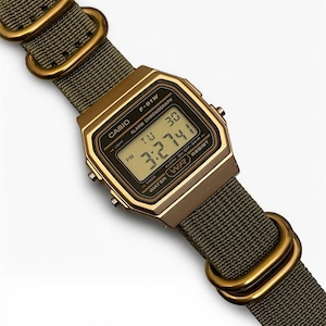 May include: A digital Casio F-91W watch with a gold-tone case and olive green nylon strap. The watch face displays the time, day, and date. The strap features gold-colored metal loops. The watch is water-resistant.
