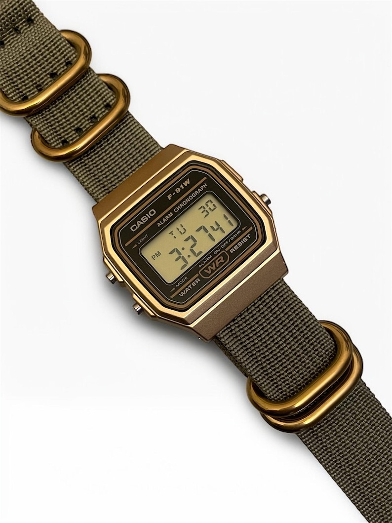 May include: A digital Casio F-91W watch with a gold-tone case and olive green nylon strap. The watch face displays the time, day, and date. The strap features gold-colored metal loops. The watch is water-resistant.