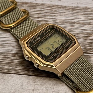 May include: A gold-toned Casio F-91W digital watch with a green nylon strap. The watch face displays the time and date. The strap features gold-colored metal loops. The watch is water resistant.