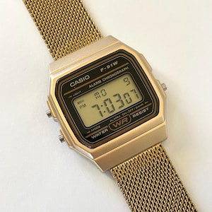 May include: A gold-toned Casio F-91W digital watch with a mesh band. The watch face displays the time, day, and date. The watch has a black digital display with the text "CASIO" and "F-91W" visible.