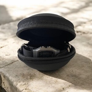 Puede incluir: Un estuche de transporte negro, semirr&iacute;gido, con cremallera y forma de concha que contiene un reloj de pulsera digital con esfera rectangular plateada y correa negra. El estuche est&aacute; entreabierto, mostrando el reloj en su interior.